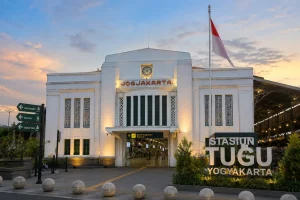 Tampak depan Stasiun Tugu Yogyakarta pada sore menjelang malam dengan fasad bangunan berwarna putih bergaya kolonial, tulisan "Stasiun Tugu Yogyakarta", bendera Merah Putih, serta langit senja yang cerah.